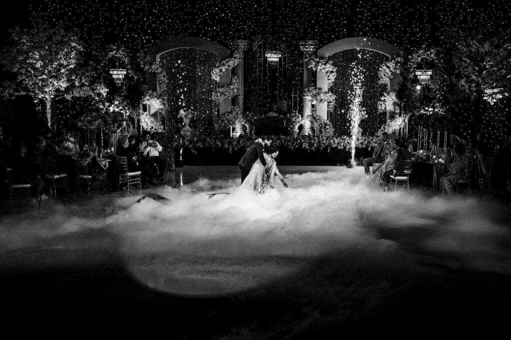 A couple dances elegantly in a mist-filled ballroom adorned with twinkling lights and floral decorations. The black and white setting creates a dreamy, romantic atmosphere, with tables and guests visible in the dimly lit background.