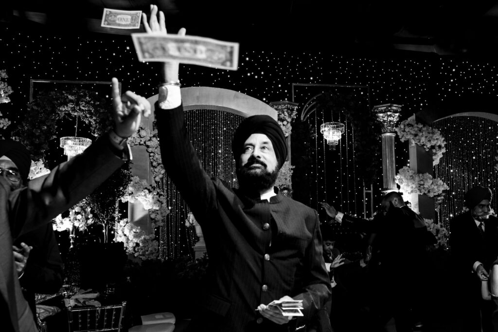 A man in a turban and suit throws money in the air at a festive event. The background is decorated with flowers and twinkling lights, creating a celebratory atmosphere. It's a black and white photo capturing the lively moment.