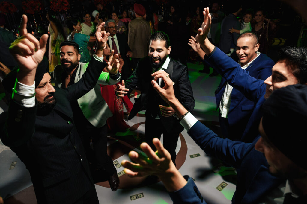 A group of men in suits are dancing energetically, surrounded by a lively crowd at an indoor event. They have their hands raised and appear to be enjoying the festive atmosphere, with colorful lights illuminating the scene.