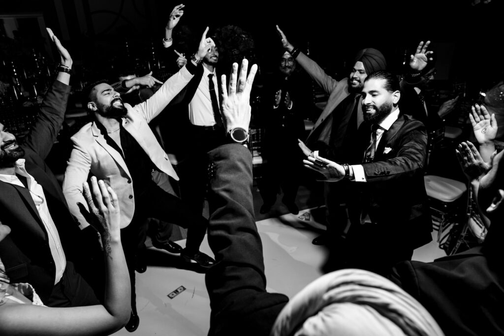 A group of men in suits joyfully dance in a circle, raising their arms. The scene is lively and upbeat, capturing a celebratory moment in a black and white photo.