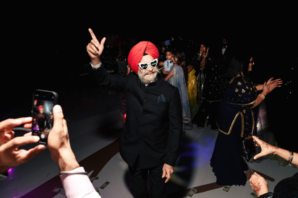 A man wearing a red turban and heart-shaped sunglasses dances on a dimly lit stage. He wears a black suit and points upwards while others, dressed in festive attire, dance around him. People take photos with their phones.