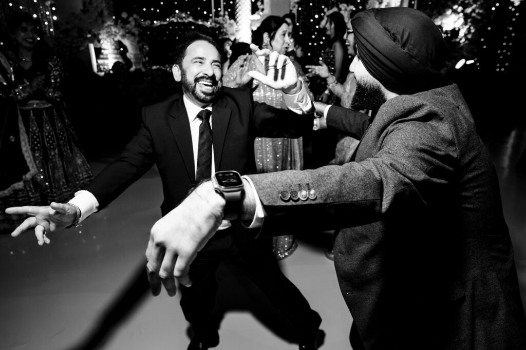 A black and white photo of two men joyfully dancing at a party. One is in a suit, and the other is wearing a turban and a jacket. The background shows festive decorations and a group of people enjoying the celebration.