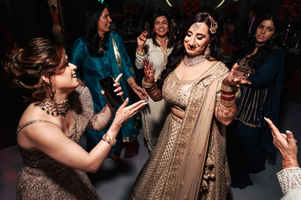 A group of women in elegant traditional attire dance joyfully at an indoor event. The focus is on one woman in a gold outfit, smiling and clapping, surrounded by friends in colorful dresses. The atmosphere is lively and festive.