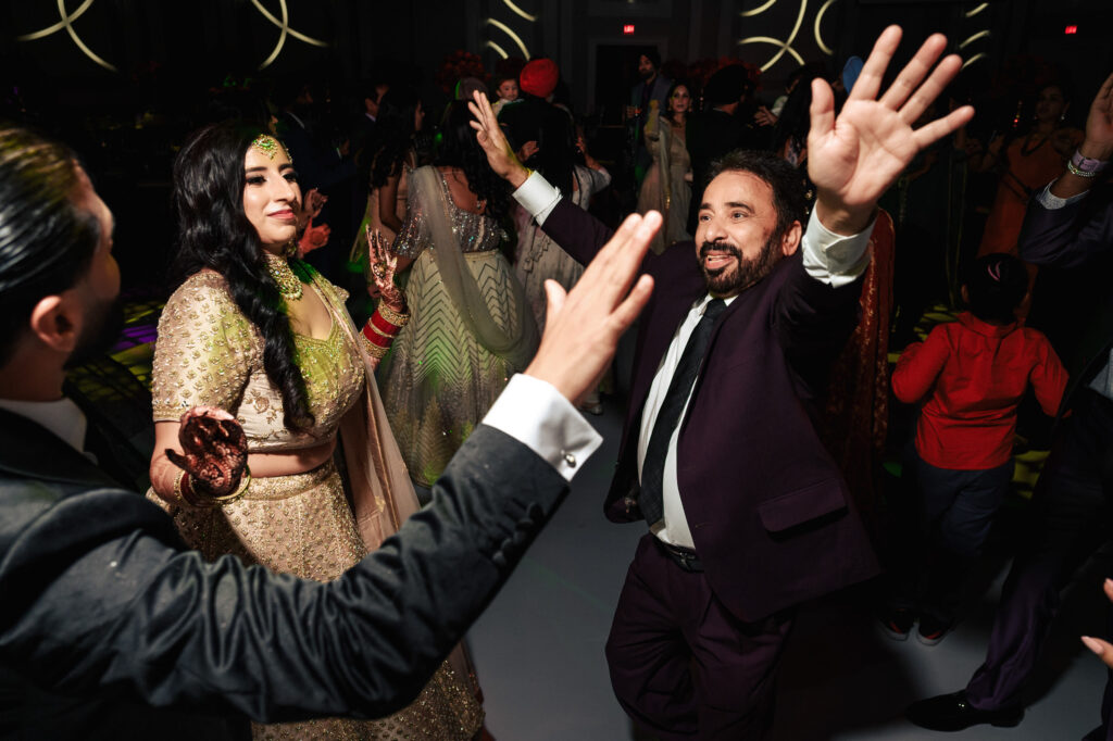 People joyfully dancing at a festive event, dressed in formal attire. A woman in a gold dress and a man in a suit are prominently celebrating under colorful lights, surrounded by other attendees.