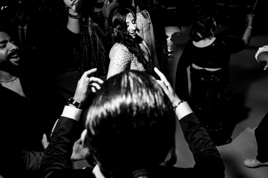 A black and white image of a joyful gathering. People are dancing, and a woman in the center is smiling while holding someone's hand. The scene is lively, with several individuals dressed in formal and traditional attire.