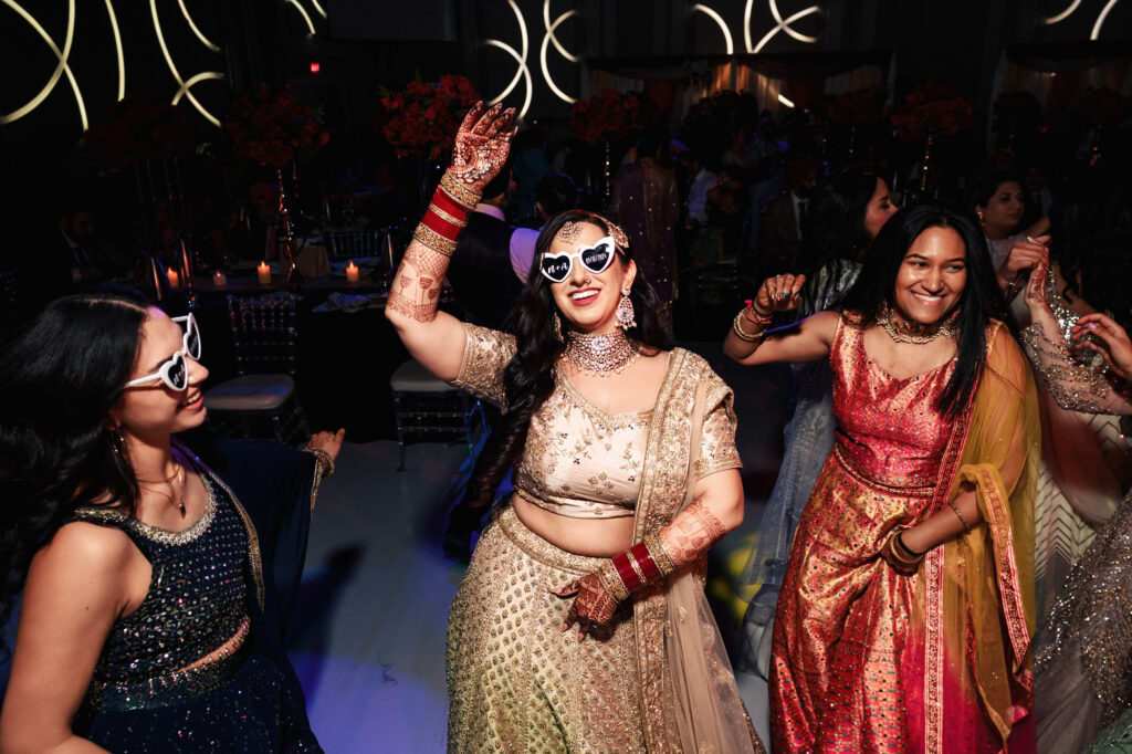 A group of women in colorful traditional Indian attire dance enthusiastically at a festive indoor celebration. The woman in the center wears heart-shaped sunglasses and intricate jewelry, enjoying the lively atmosphere.