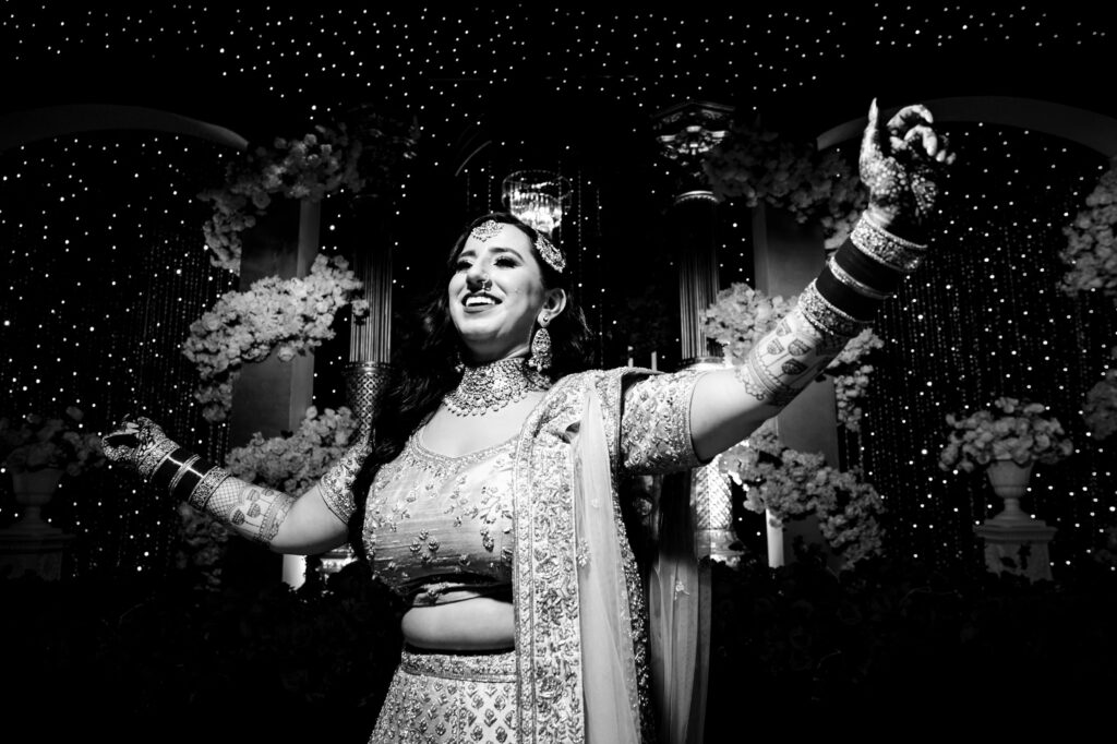 A woman dressed in traditional attire joyfully dances with her arms raised. The background is adorned with sparkling lights and floral decorations, creating a festive ambiance. The image is in black and white.