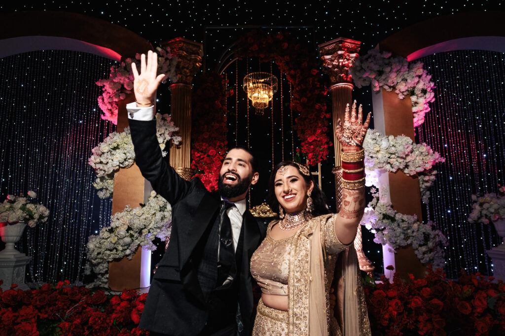 A joyous couple in formal attire waves amid a lavish setting with abundant floral arrangements and twinkling lights. The woman wears a traditional embellished outfit with henna on her hands, while the man is in a suit.