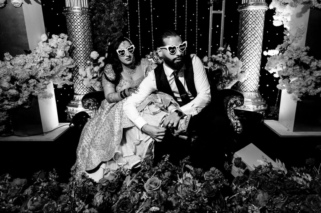 A couple dressed in formal attire sits on a lavishly decorated couch. They both wear heart-shaped sunglasses. The background features ornate columns and floral arrangements, adding to the festive ambiance.