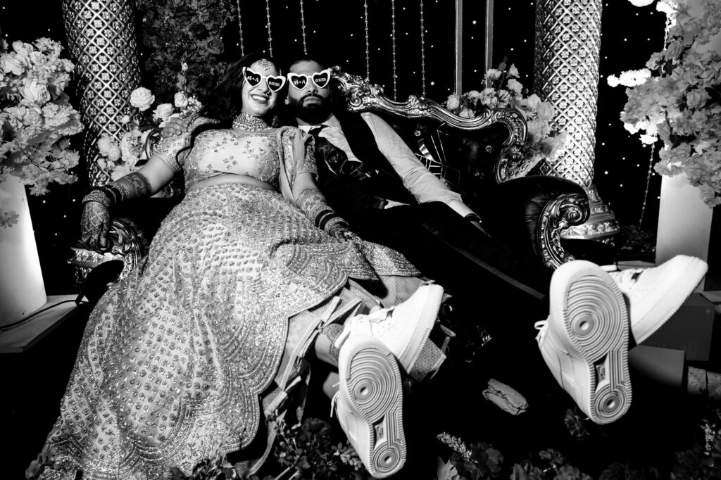 A couple wearing heart-shaped sunglasses relaxes on an ornate couch at a wedding. The scene is decorated with flowers and lights. They wear traditional attire and sneakers, smiling and enjoying the celebration. The image is in black and white.