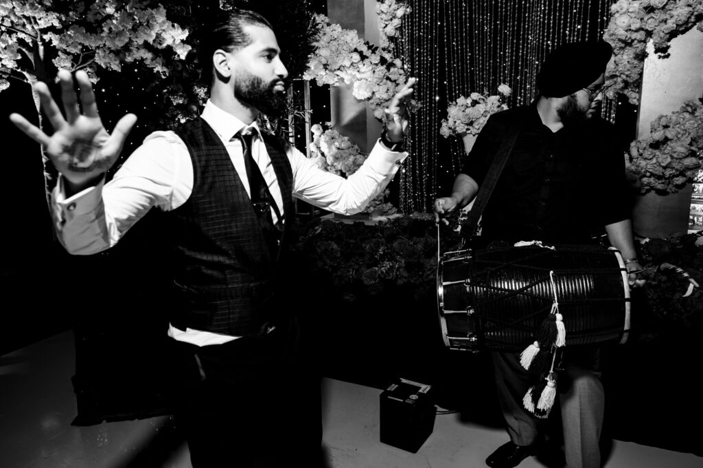 A black and white photo of a man in a vest and tie, gesturing with his hands, while another man in a turban plays a dhol drum. They are surrounded by floral decorations and string lights.