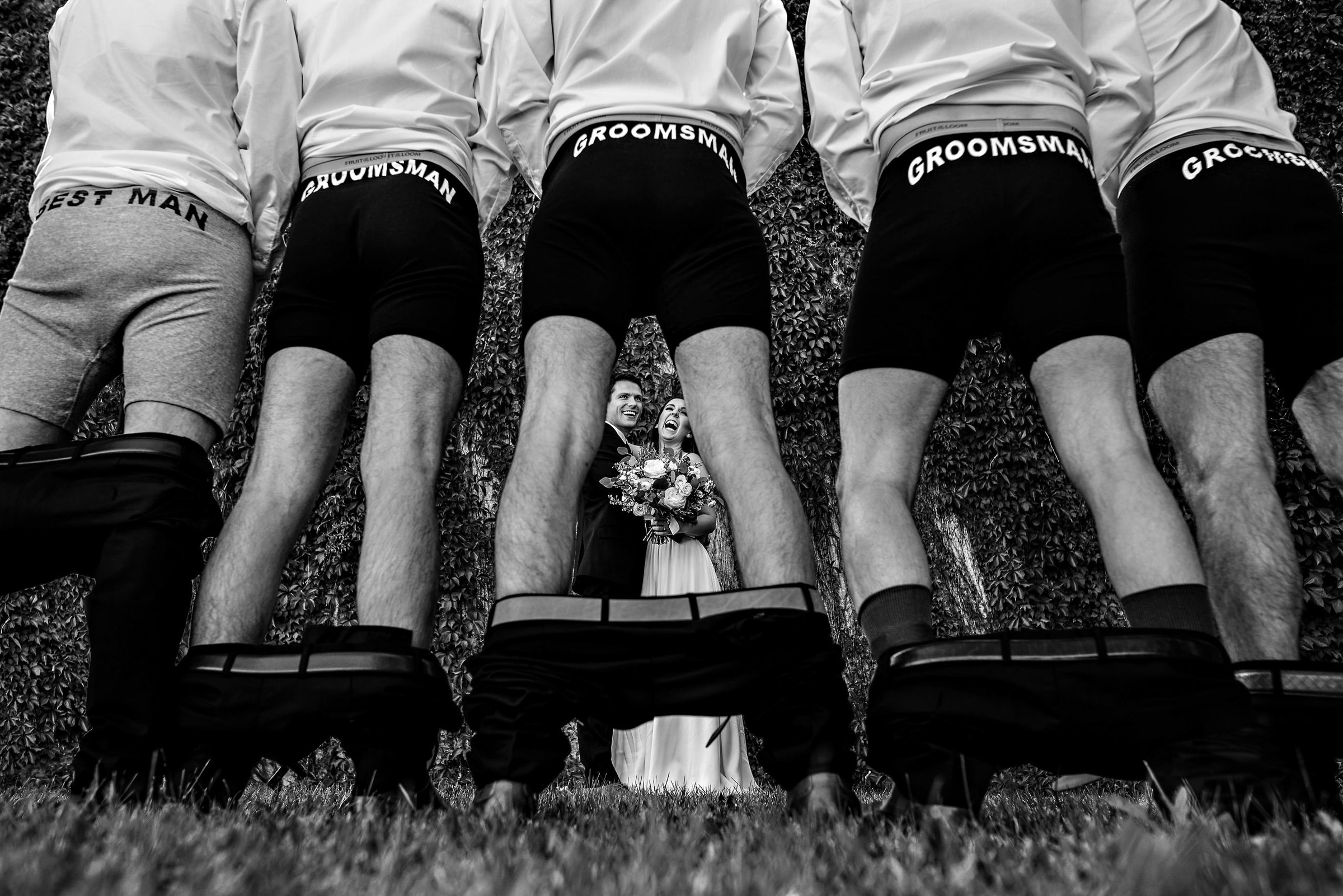 A black-and-white photo by a talented Winnipeg wedding photographer captures a bride and groom beaming as they pose. In the foreground, four groomsmen face away, wearing shirts and boxer briefs labeled "Groomsman" and "Best Man," with their pants cheekily pulled down.
