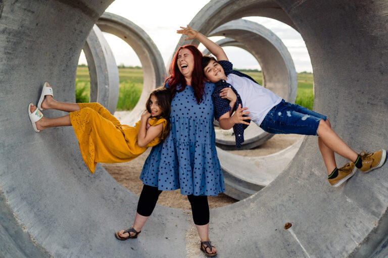 A Winnipeg photographer captures a woman playfully standing in large concrete pipe structures, holding two children. The girl on her left wears a yellow dress, and the boy on her right sports denim shorts. Their joy is palpable against the vibrant outdoor backdrop.