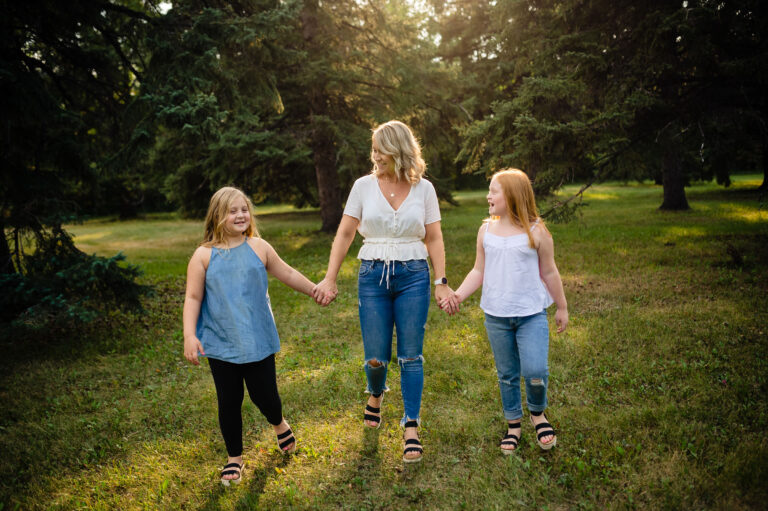 A woman and two girls hold hands, smiling as they stroll through a sunlit, grassy area surrounded by trees. The woman, in a white top and jeans, stands between the girls in their casual attire. Captured beautifully by a Winnipeg photographer, this moment radiates joy and warmth.