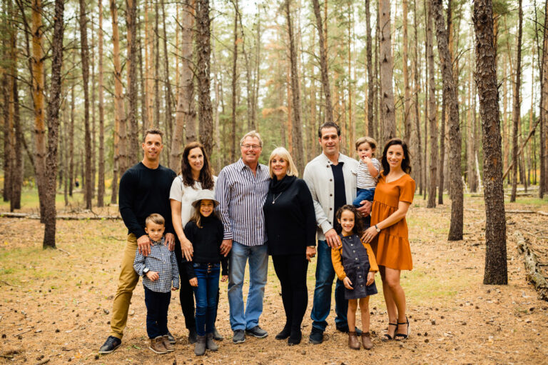 A family of nine, captured by a talented Winnipeg photographer, poses in a wooded area with tall trees. The group includes four adults, two young children, two older children, and a baby. Dressed in casual fall attire, they smile warmly at the camera amidst the fallen leaves.