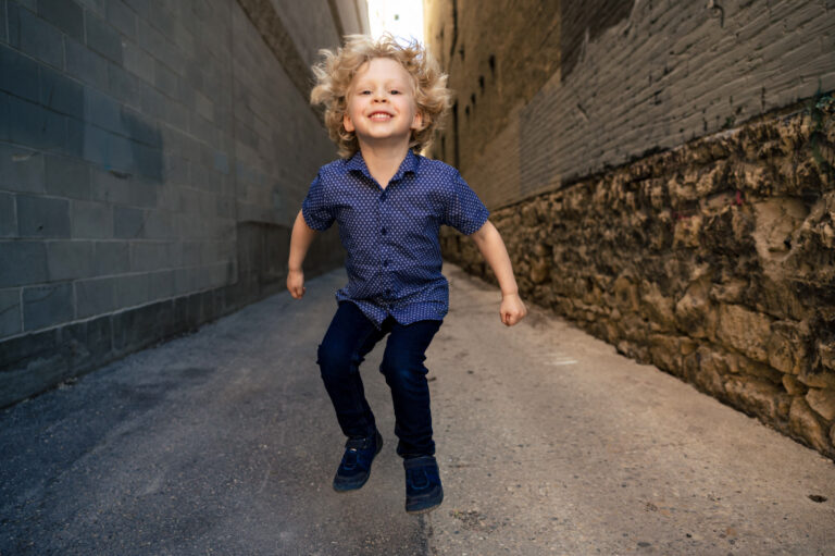 A happy child with curly blonde hair is joyfully jumping in a narrow alleyway lined with brick and stone walls, captured perfectly by a Winnipeg photographer. The child wears a blue patterned shirt and dark pants, beaming with an infectious smile.