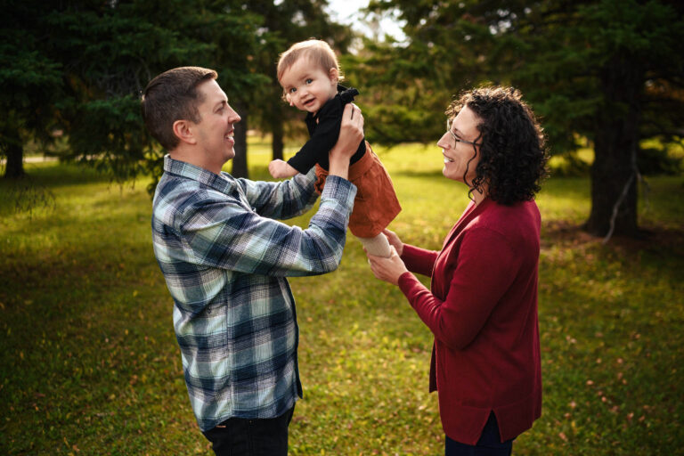A Winnipeg photographer captures a joyous moment as a man in a plaid shirt lifts a smiling baby in a black top and rust-colored skirt. A woman in a red cardigan leans in from the side, smiling brightly. They stand on a grassy area with lush green trees in the background.