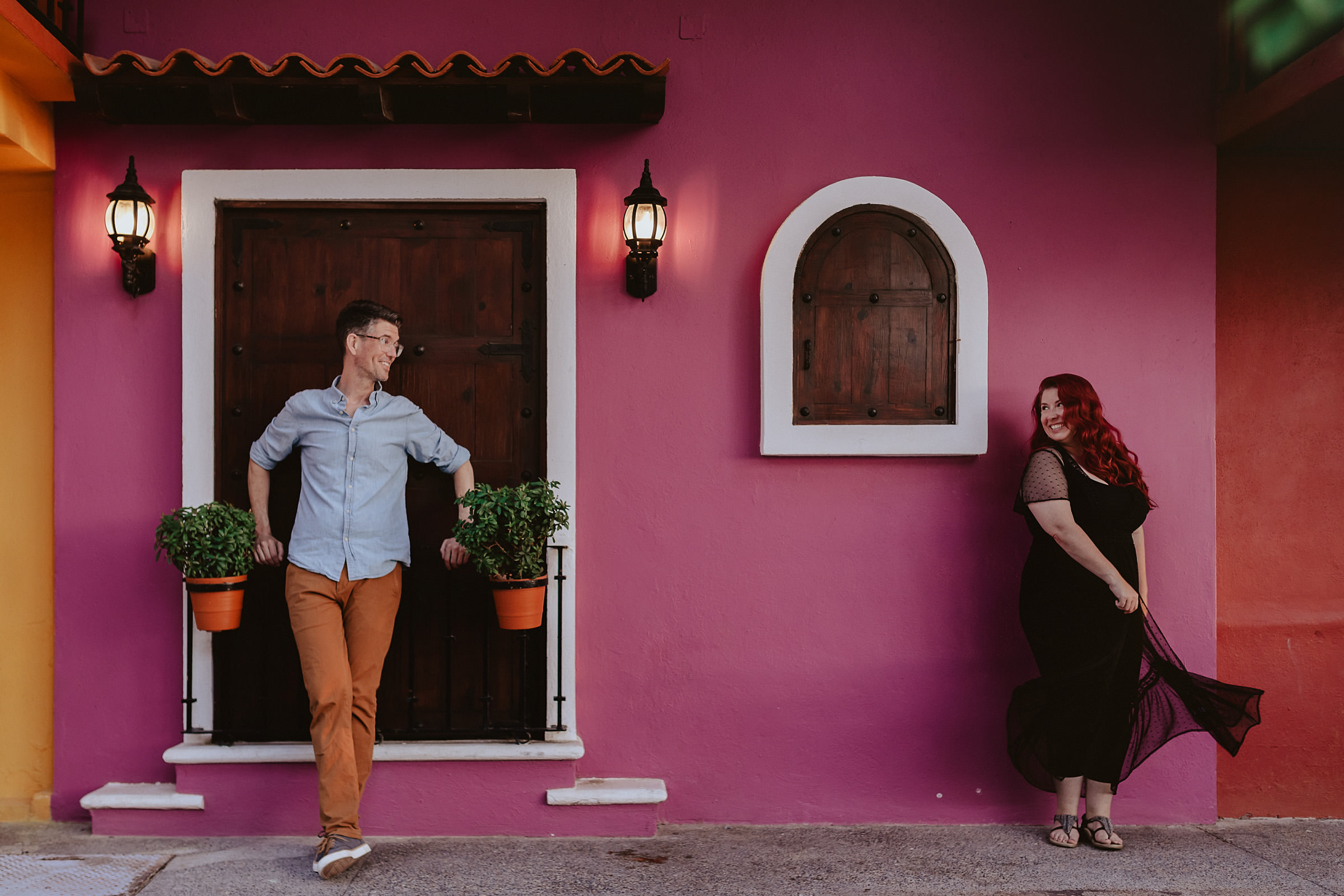 A man leans against a pink wall with a wooden door, smiling at a woman in a black dress. She's standing by a small arched window. Captured by a Winnipeg wedding photographer, the scene is adorned with two potted plants and illuminated by outdoor lanterns.