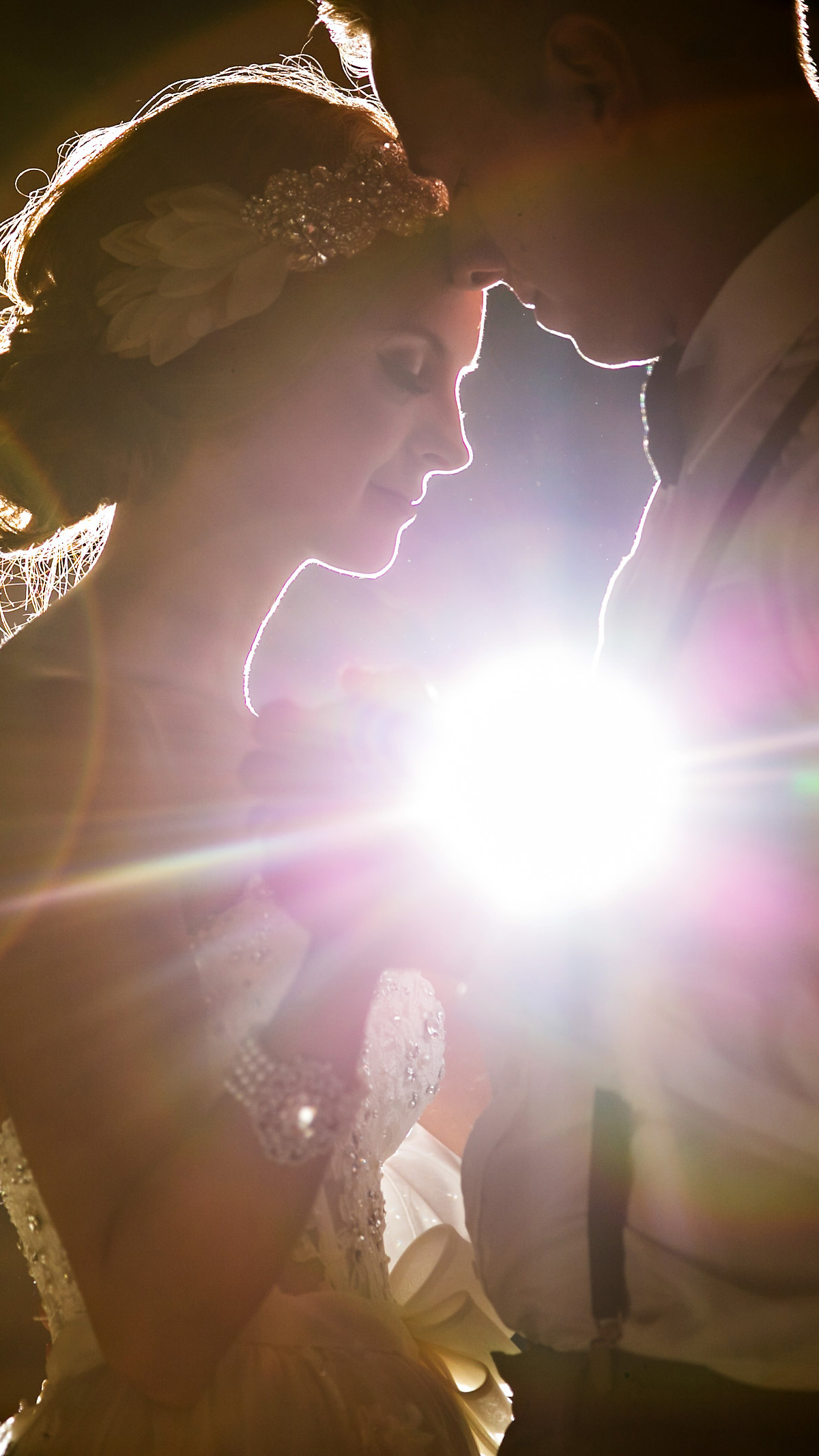 A couple dances closely, silhouetted by bright light as captured by a Winnipeg wedding photographer. She's adorned with a headpiece and gown, while he's in a white shirt with suspenders, their faces gently touching.