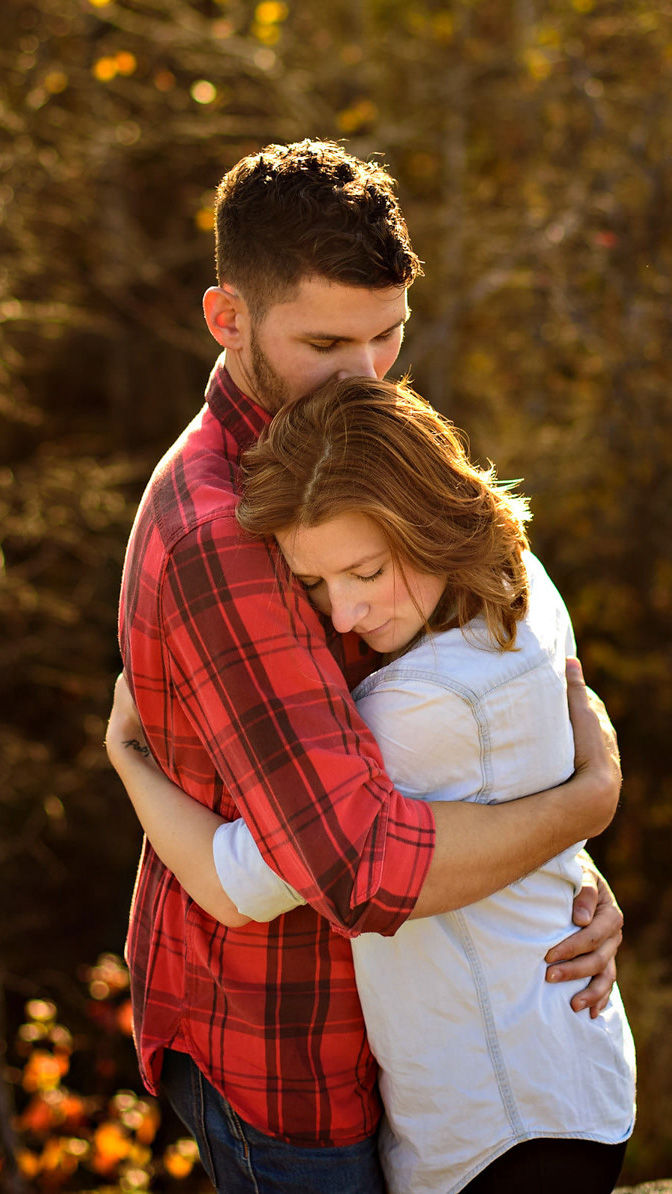 A couple standing outdoors share a tender embrace, captured beautifully by a Winnipeg wedding photographer. The man, in a red plaid shirt, gently holds the woman in a light top as she rests her head on his shoulder. The softly blurred background features the warm hues of autumn foliage.