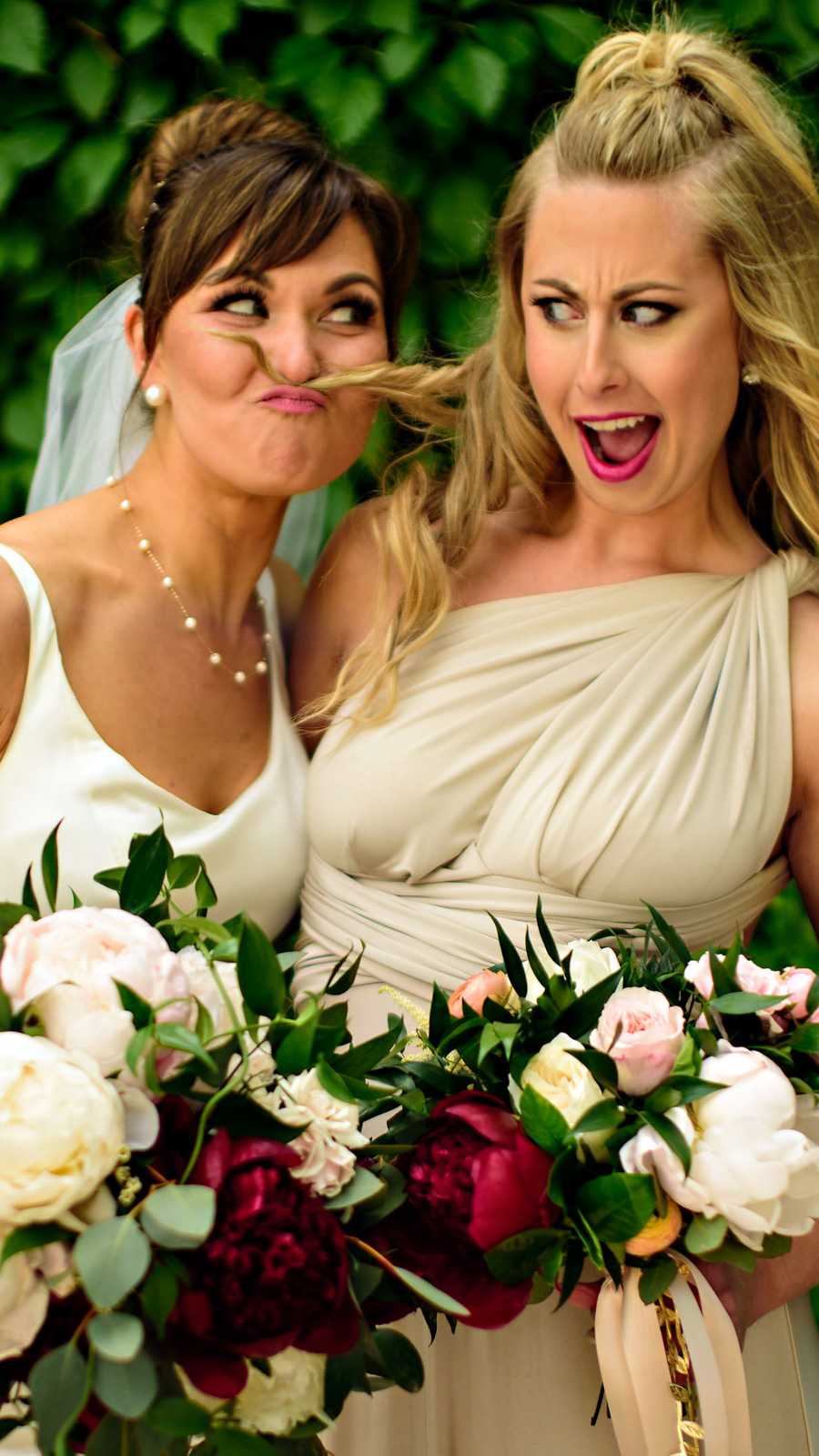 Two women in white dresses make playful faces, holding bouquets of red and white flowers. The woman on the left sports a veil and uses her hair to create a mustache, while the one on the right looks surprised. Captured by a Winnipeg wedding photographer, lush greenery serves as their backdrop.