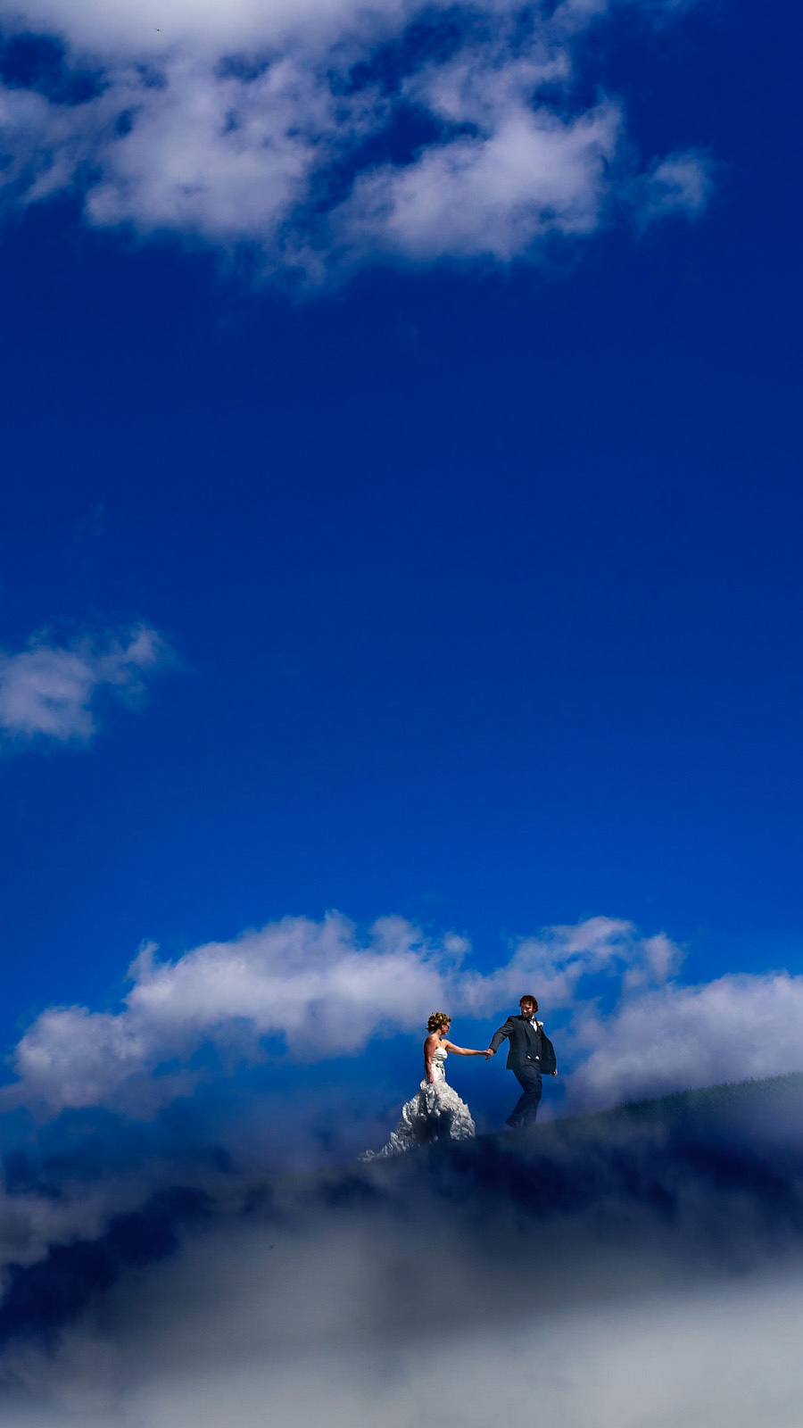 A bride and groom hold hands atop a cloud-like formation, captured beautifully by a Winnipeg wedding photographer. The vast blue sky is dotted with fluffy white clouds, creating a surreal and romantic atmosphere.