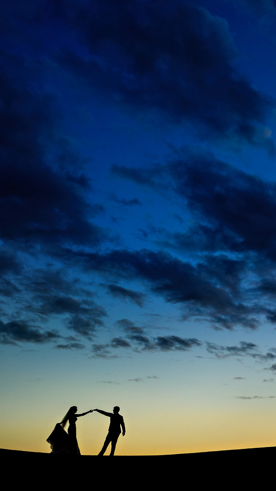 Silhouetted couple dancing against a dramatic sky at sunset, with shades of blue and orange. The scene, beautifully captured by a Winnipeg wedding photographer, showcases scattered clouds creating a picturesque and romantic atmosphere.