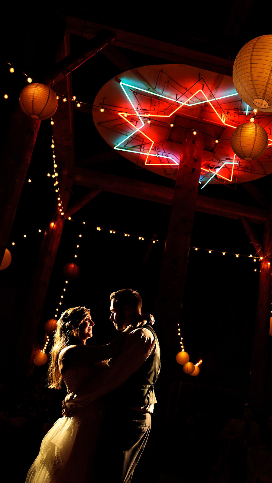 A couple dances closely under dim lights and a large neon star in a cozy setting, perfectly capturing the essence that any Winnipeg Wedding Photographer would treasure. Warm string lights and spherical paper lanterns add to the romantic atmosphere as she twirls in her white dress beside him in his dark suit.