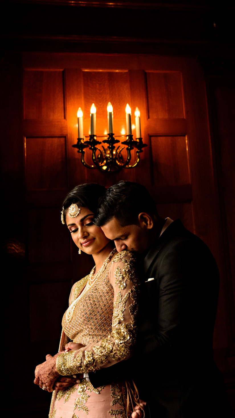 A couple embraces in front of a wooden paneled wall illuminated by a candle-style chandelier. Captured by a Winnipeg wedding photographer, the woman wears a detailed, patterned dress with jewelry, while the man in a suit affectionately hugs her from behind, both exuding warmth and intimacy.