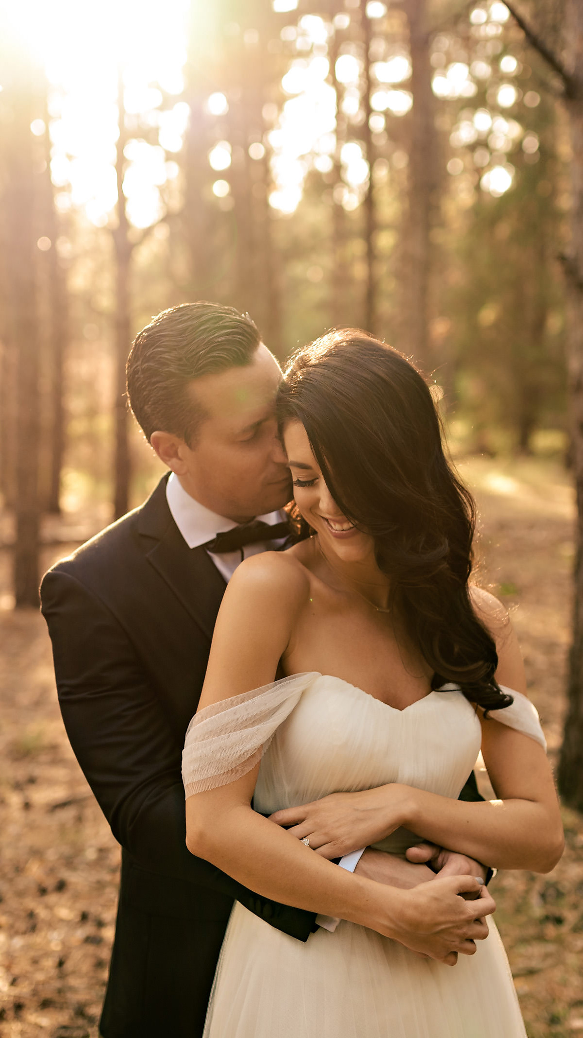 A bride and groom, captured by a Winnipeg wedding photographer, embrace in a sunlit forest. The groom, in a dark suit, gently kisses the bride's head as she smiles in her off-shoulder white dress. Warm sunlight filters through the trees, creating a romantic atmosphere.