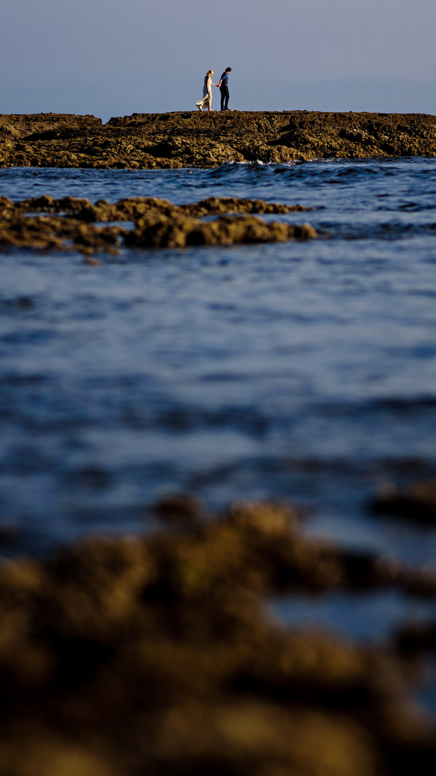 A couple strolls along a rugged, rocky shoreline beneath a clear blue sky. In the foreground, ocean waves gently crash against the rocks, captured beautifully through the lens of a Winnipeg wedding photographer, preserving their moments in timeless elegance.