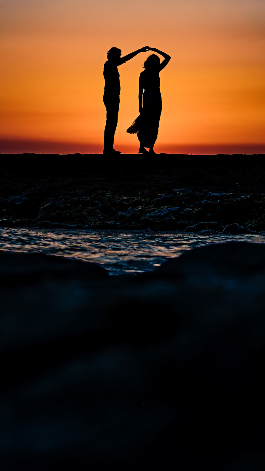 A silhouetted couple dances on a rocky shore during a vibrant sunset, captured beautifully by a Winnipeg wedding photographer. The sky transitions from orange to blue, with gentle waves in the foreground, softly highlighting the couple's figures against the horizon.