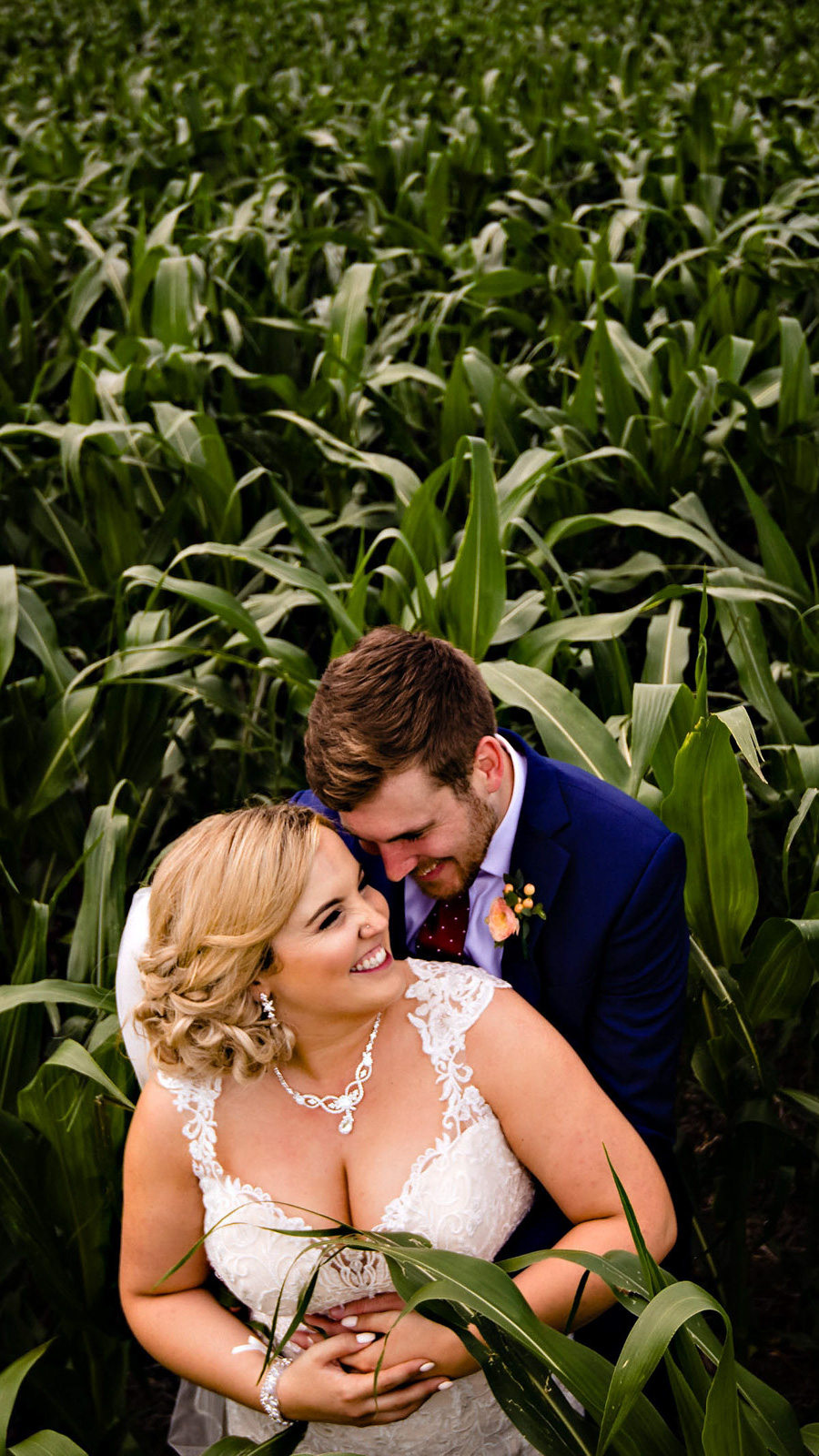 A couple embraces in a lush cornfield, captured beautifully by a Winnipeg wedding photographer. The woman, in a white lace dress, smiles as she looks up. The man, in a navy suit and purple tie, stands behind her affectionately. Both appear joyful and relaxed.