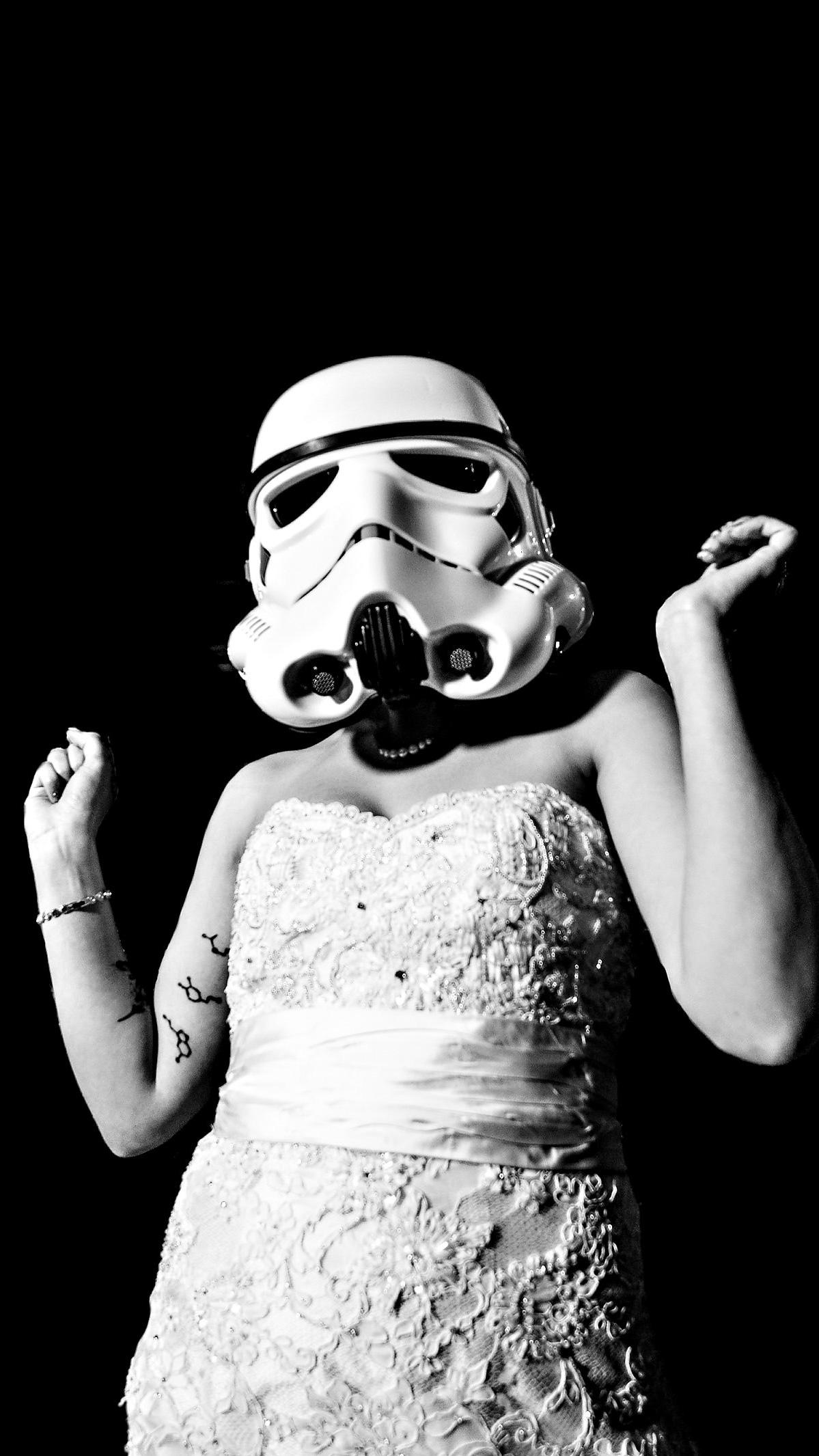 A person wearing a Star Wars stormtrooper helmet poses elegantly in a strapless, lace wedding dress adorned with decorative beads. Their arms reveal subtle tattoos. Captured by a Winnipeg wedding photographer, the black backdrop enhances the dramatic contrast with the white attire.