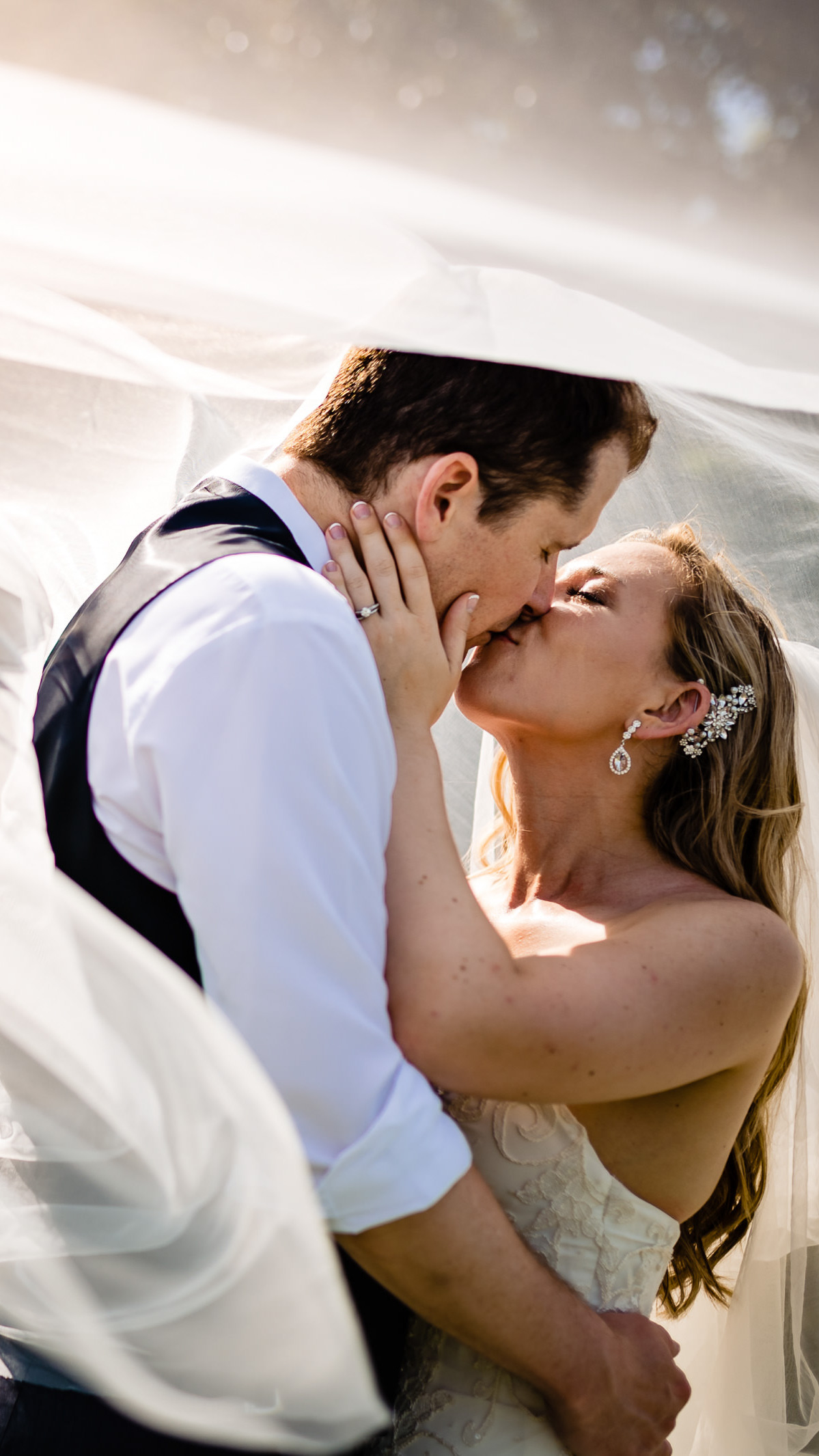 A bride and groom share a kiss under a flowing veil, beautifully captured by a Winnipeg wedding photographer. The bride, in her strapless dress and jeweled hairpiece, embraces the groom in his white shirt and dark vest. Sunlight filters through, enhancing the soft, intimate atmosphere.