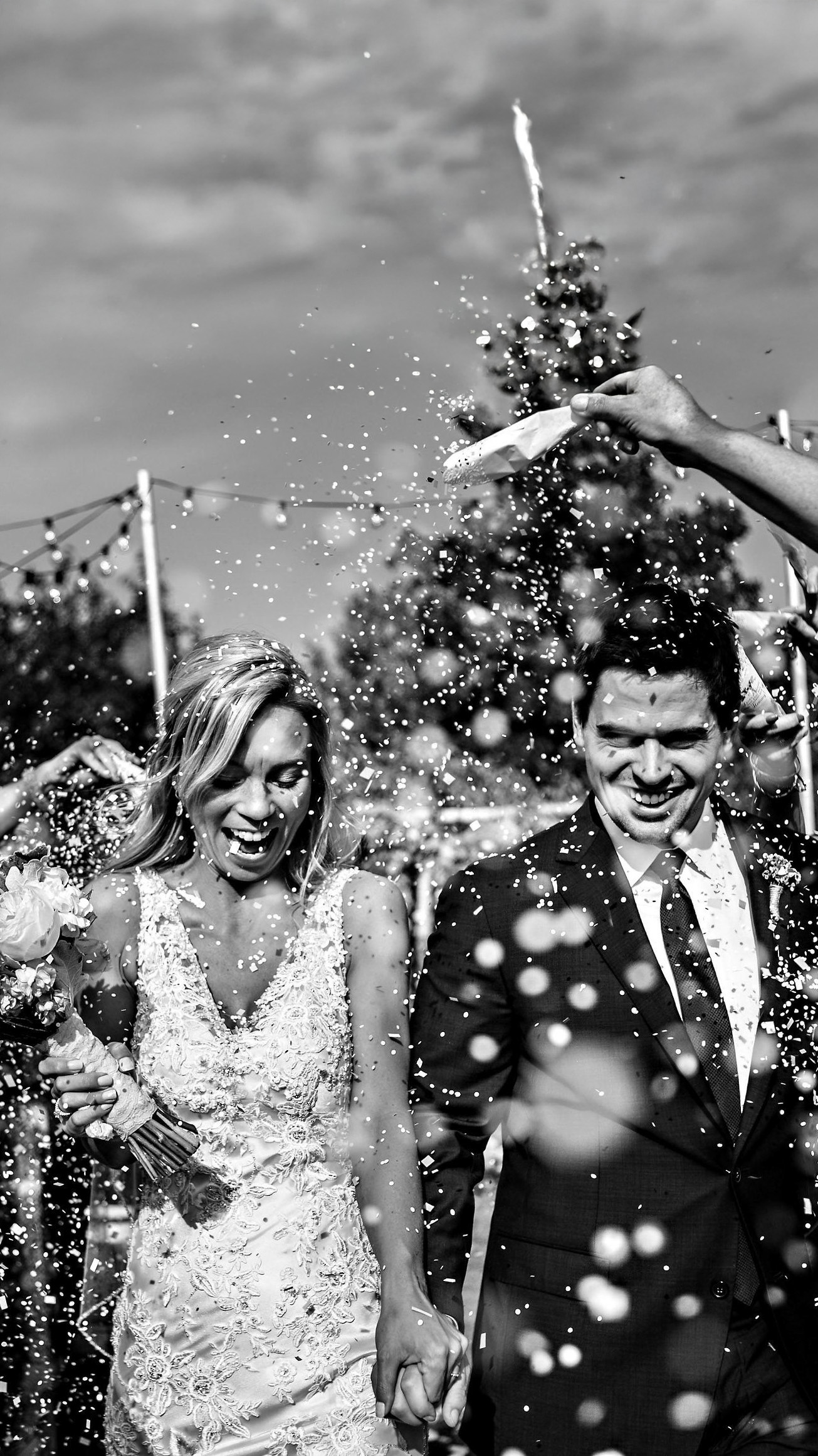 A joyous bride in a lace dress and a groom in a suit are captured by a Winnipeg wedding photographer as they walk outdoors, guests showering them with confetti. The couple beams, holding hands amid festive decorations under the clear sky.
