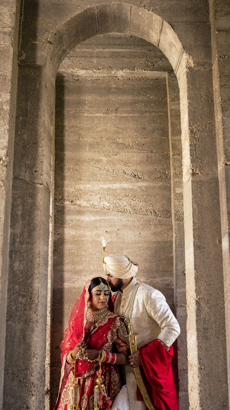 A couple in traditional Indian wedding attire poses gracefully under a stone archway, captured beautifully by a skilled Winnipeg wedding photographer. The bride dazzles in a red and gold lehenga as the groom, in his white sherwani with a red turban, leans in to kiss her forehead tenderly.