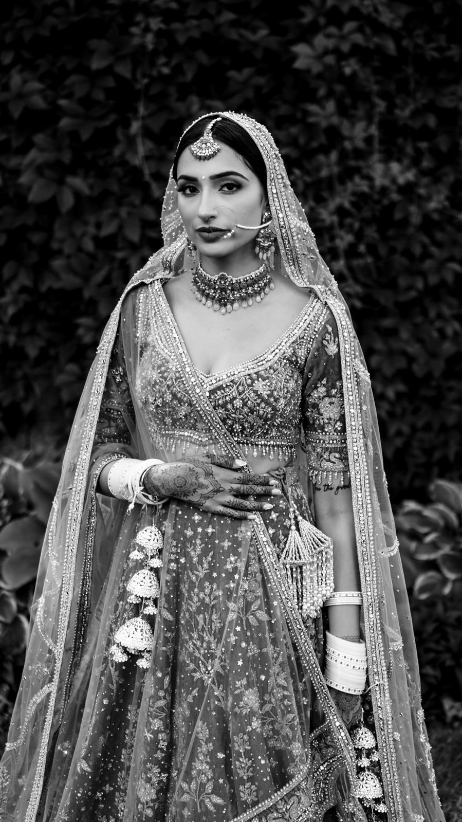 A woman in a detailed, traditional outfit stands against a leafy backdrop. She wears a richly embroidered dress with a matching dupatta, intricate jewelry, and henna on her hands. Captured by a Winnipeg wedding photographer, the black and white image is timelessly elegant.