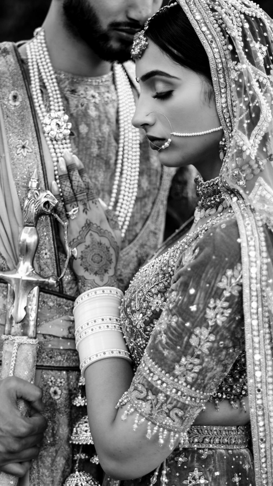 A bride and groom in traditional attire stand closely, captured by a Winnipeg wedding photographer. The bride, adorned with detailed embroidery, jewelry, and henna on her hands, looks downwards while the groom holds a decorative sword. The image is beautifully rendered in black and white.