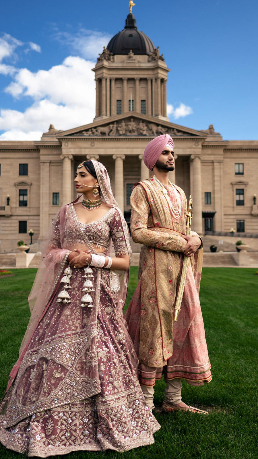 A couple stands in front of a grand building with a dome, captured beautifully by a Winnipeg wedding photographer. They're wearing traditional South Asian wedding attire in shades of pink and gold. The woman dazzles in an ornate lehenga, while the man complements her in a matching sherwani and turban under the clear sky.