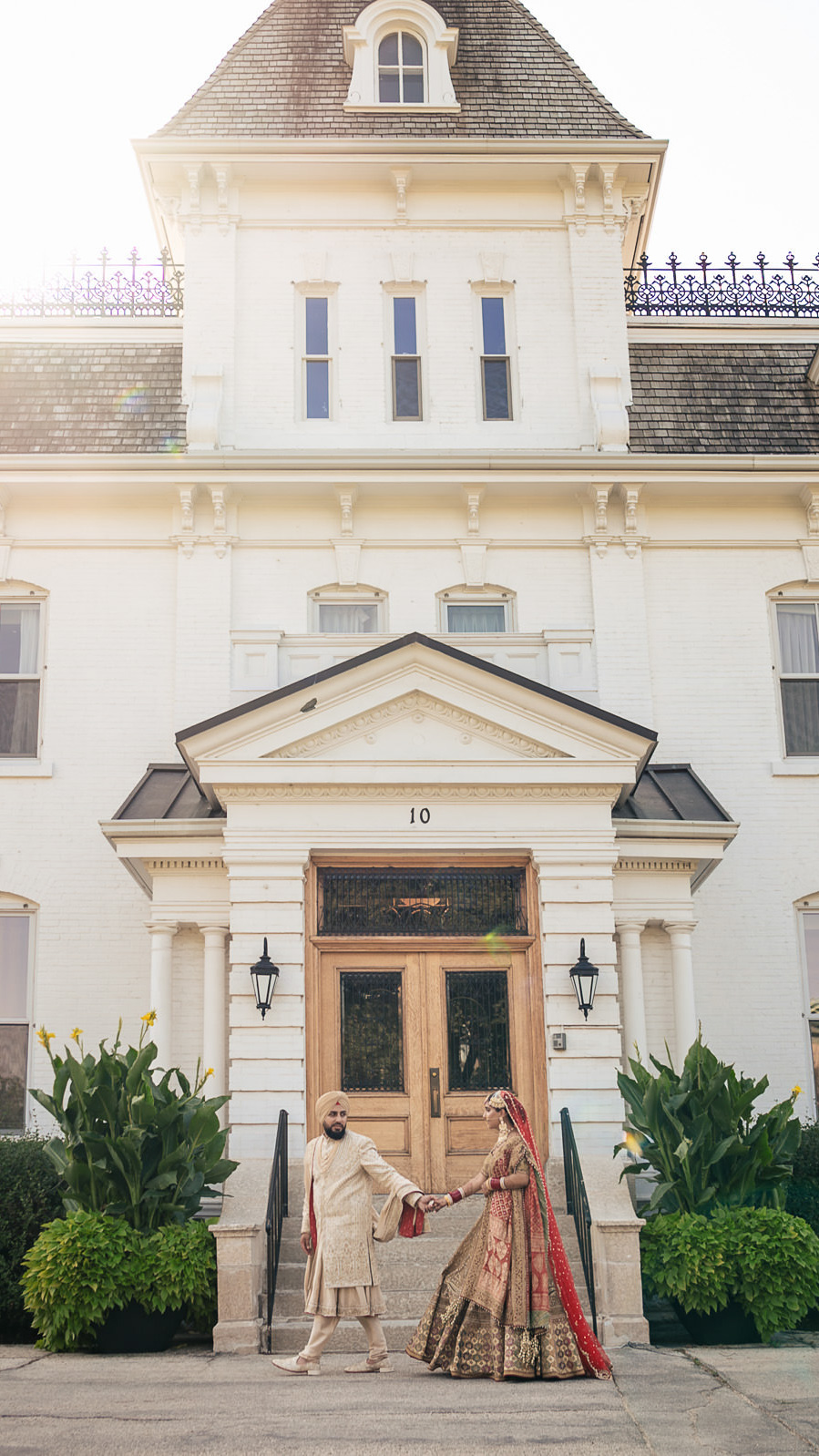 A couple in traditional South Asian attire poses elegantly before a grand building. The man, in a beige sherwani and turban, and the woman, in a red and gold bridal lehenga, are beautifully captured by a Winnipeg wedding photographer. Lush greenery adorns the scene as sunlight streams from the top left.