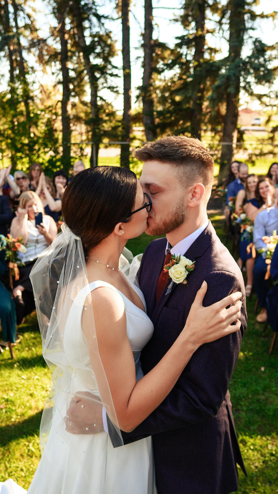 A bride and groom share a kiss outdoors, surrounded by trees and seated guests, as their Winnipeg wedding photographer captures the moment. The bride wears a white gown and veil, while the groom is in a dark suit with a boutonniere. Sunlight filters through the trees, creating a warm atmosphere.