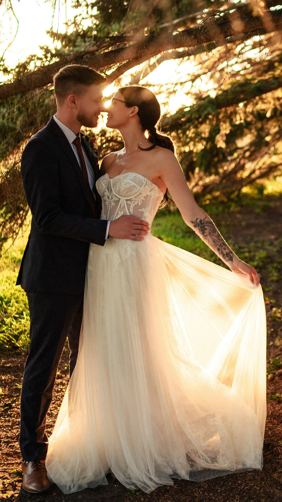 In a serene forest setting, captured by a Winnipeg wedding photographer, a couple embraces tenderly. The bride's flowing white gown with lace details complements the groom's dark suit as sunlight filters through the trees, casting a warm glow on their intimate moment.