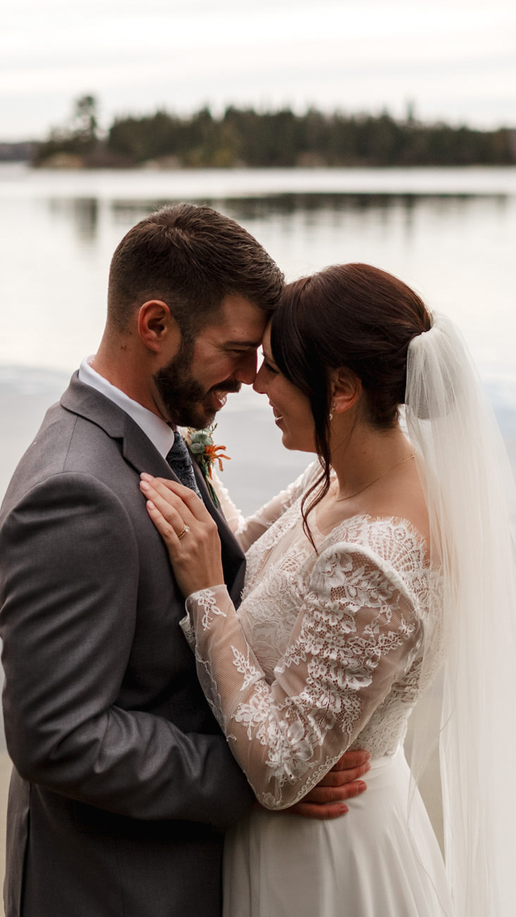 Captured by a Winnipeg Wedding Photographer, the bride and groom embrace closely by a calm lakeside. She wears a lace wedding dress with a veil, and he dons a gray suit with a boutonniere. The serene lake and distant trees complete this picturesque moment.