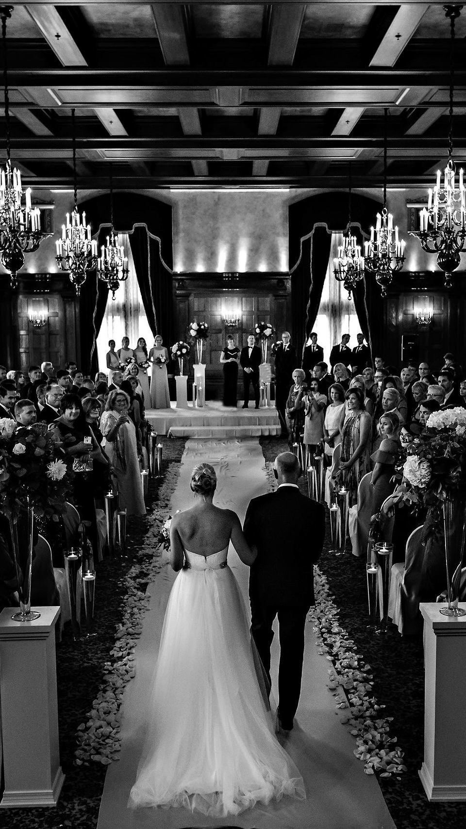A black and white image captures a Winnipeg Wedding Photographer's vision of a wedding ceremony in an elegant hall with chandeliers. The bride and groom walk down a petal-strewn aisle flanked by guests, while bridesmaids and groomsmen stand at the front against dark draped curtains.
