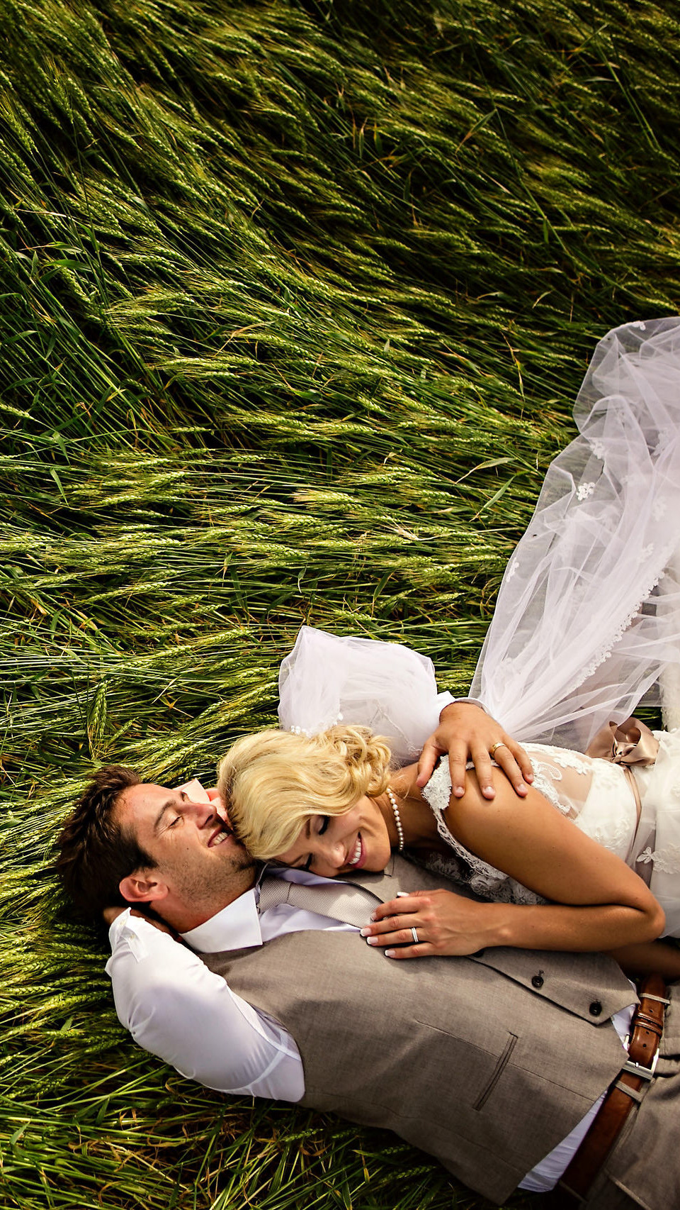A couple lies on lush green grass, smiling and embracing. The woman, in a white wedding dress, rests her head on the man's chest. Captured by a skilled Winnipeg wedding photographer, the man wears a white shirt and gray vest with eyes closed. They appear content and relaxed in a field.