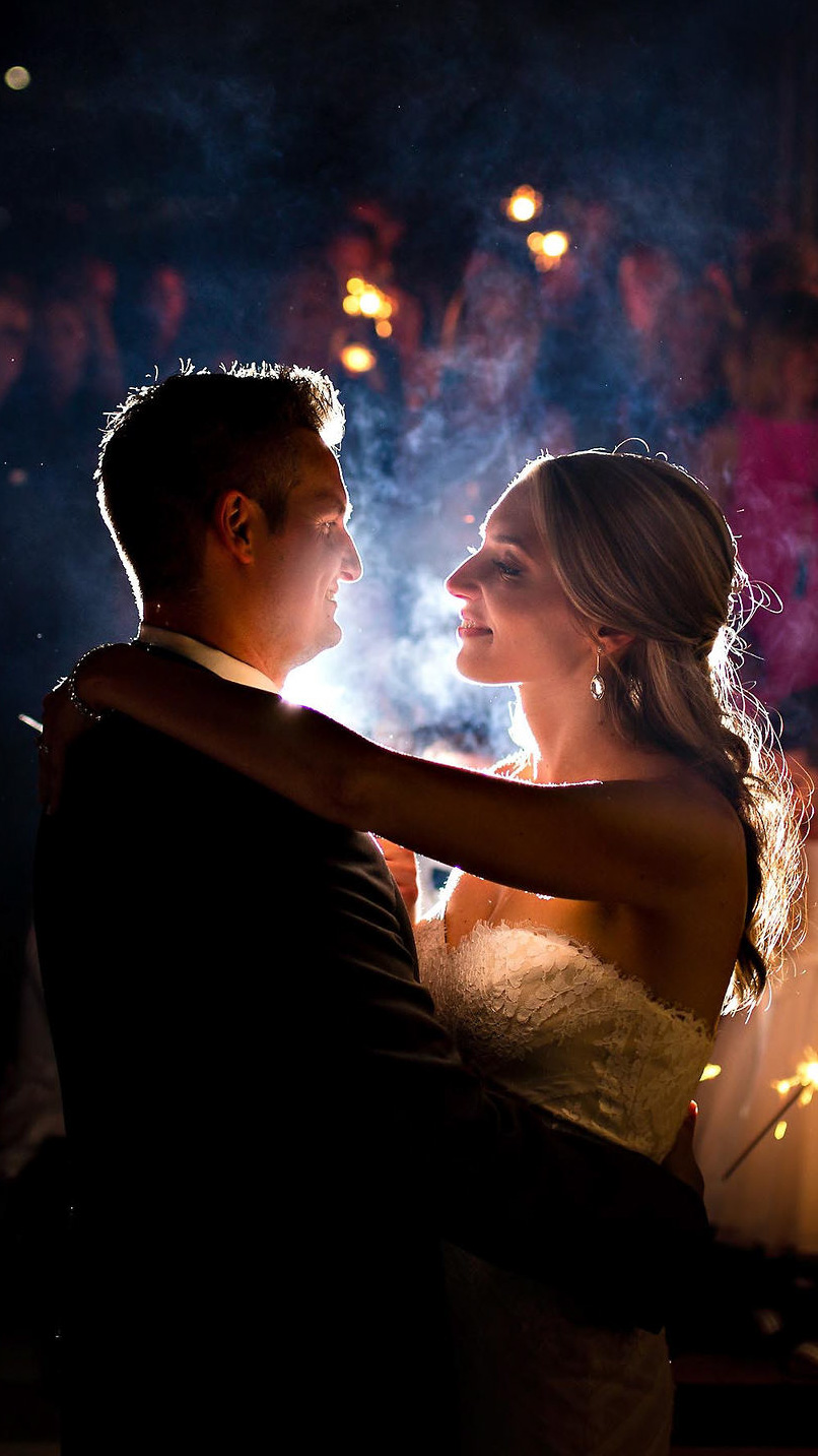 A bride and groom share a dance, illuminated by soft light. The bride wears a white dress and the groom is in a suit. They gaze at each other lovingly, captured perfectly by their Winnipeg wedding photographer, surrounded by dimly lit figures holding sparklers in the background.