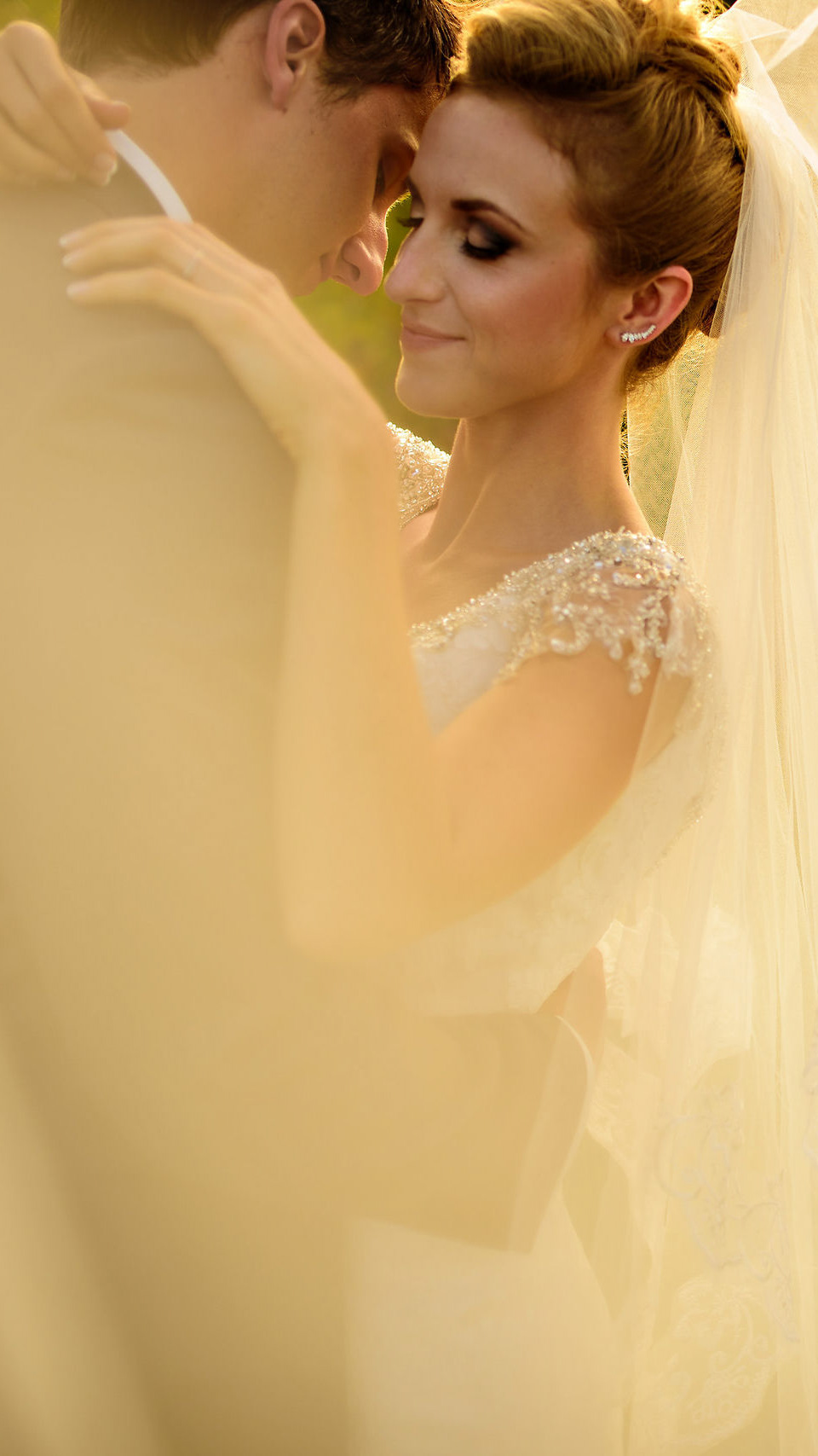 A bride and groom embrace closely, with soft light creating a dreamy effect captured by a talented Winnipeg Wedding Photographer. The bride is in a lacy white dress with a veil, and the groom wears a suit. The scene conveys a sense of romance and warmth.