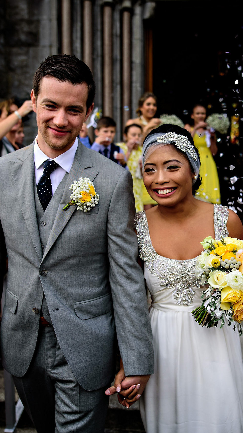 A bride and groom smile joyfully while walking hand in hand, captured perfectly by a Winnipeg wedding photographer. The groom wears a gray suit with a floral boutonniere, and the bride is in a white dress holding a yellow and white bouquet. Guests in the background are celebrating with confetti.