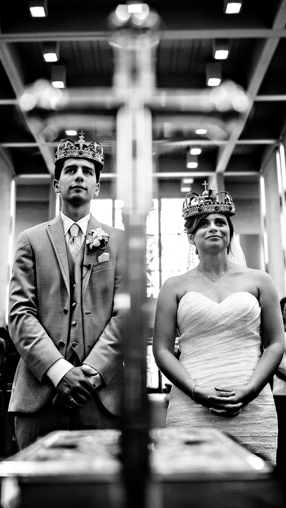 A bride and groom, both wearing crowns, stand inside a church, captured by a Winnipeg wedding photographer. The groom is in a suit with a boutonniere, and the bride is in a strapless gown with a veil. They are positioned on either side of a blurred cross in the foreground—black and white photo.